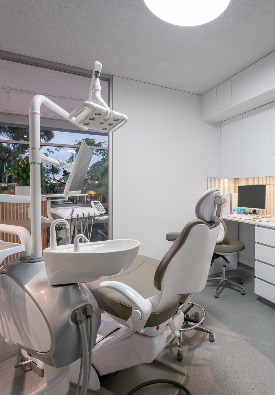Modern dental treatment room with a patient chair, white cabinetry, and large windows.