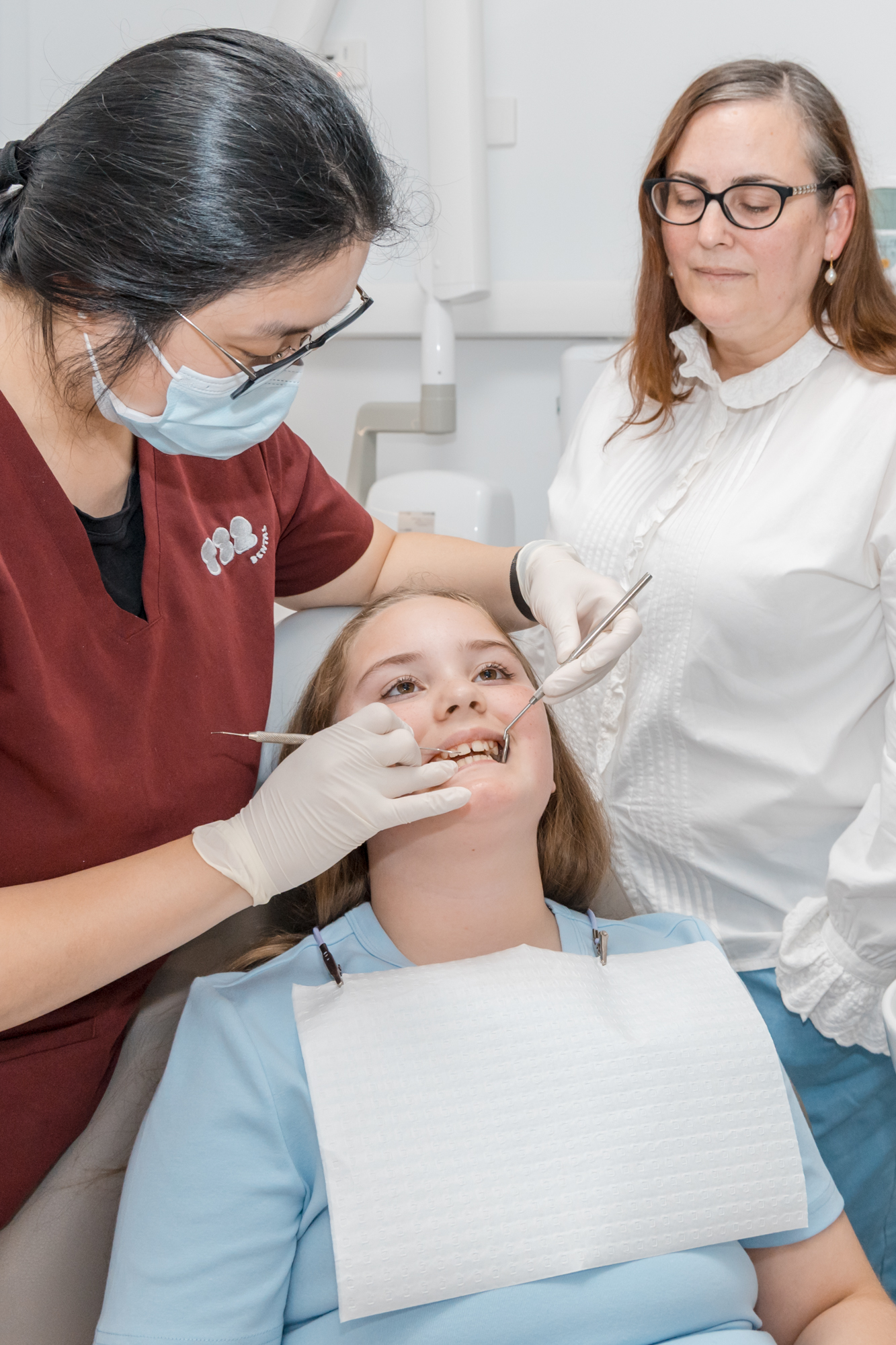Dentist examining a young patient's teeth with clinical tools while an adult woman watches.
