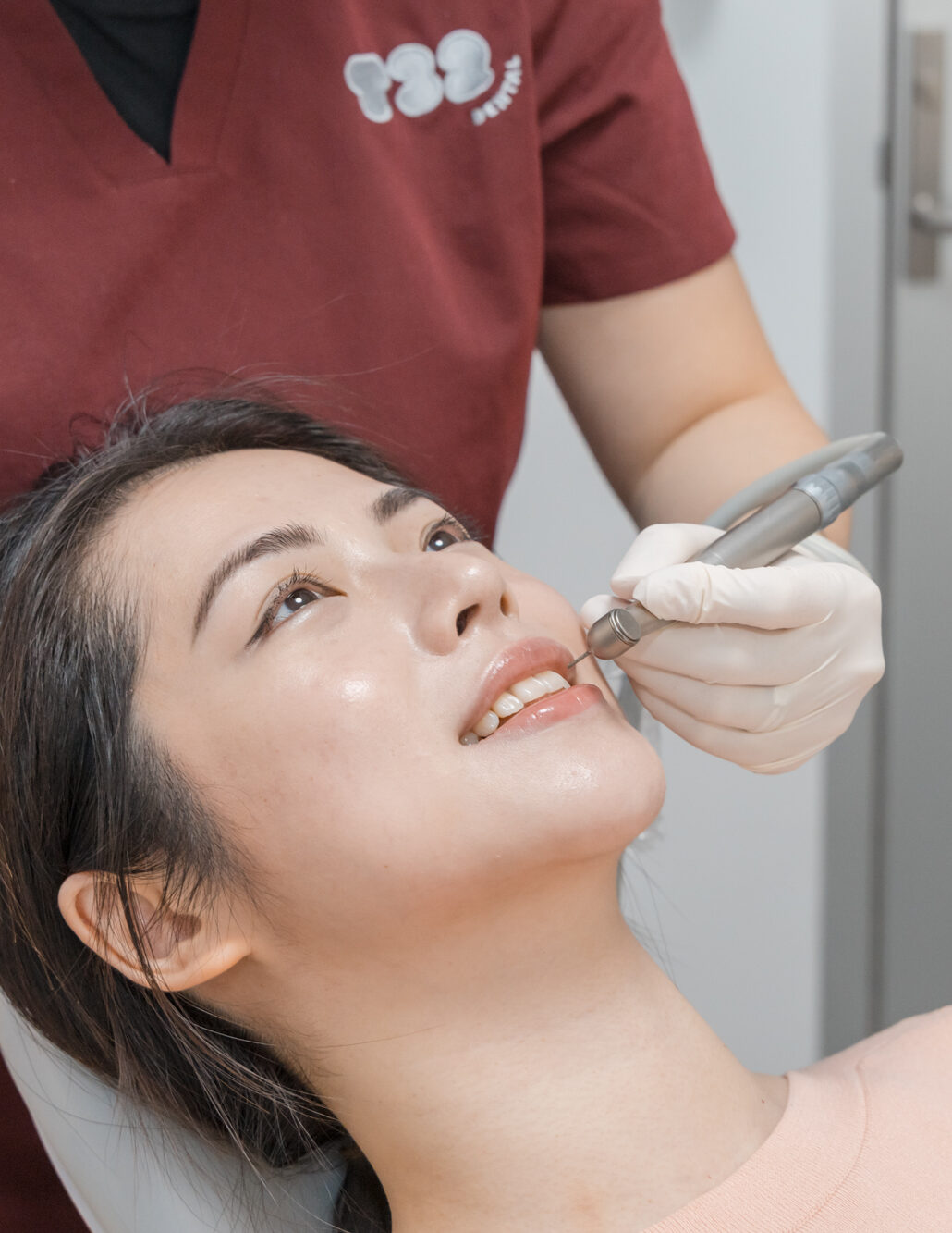 Dentist using a handpiece drill on a smiling female patient's teeth during a clinical appointment.