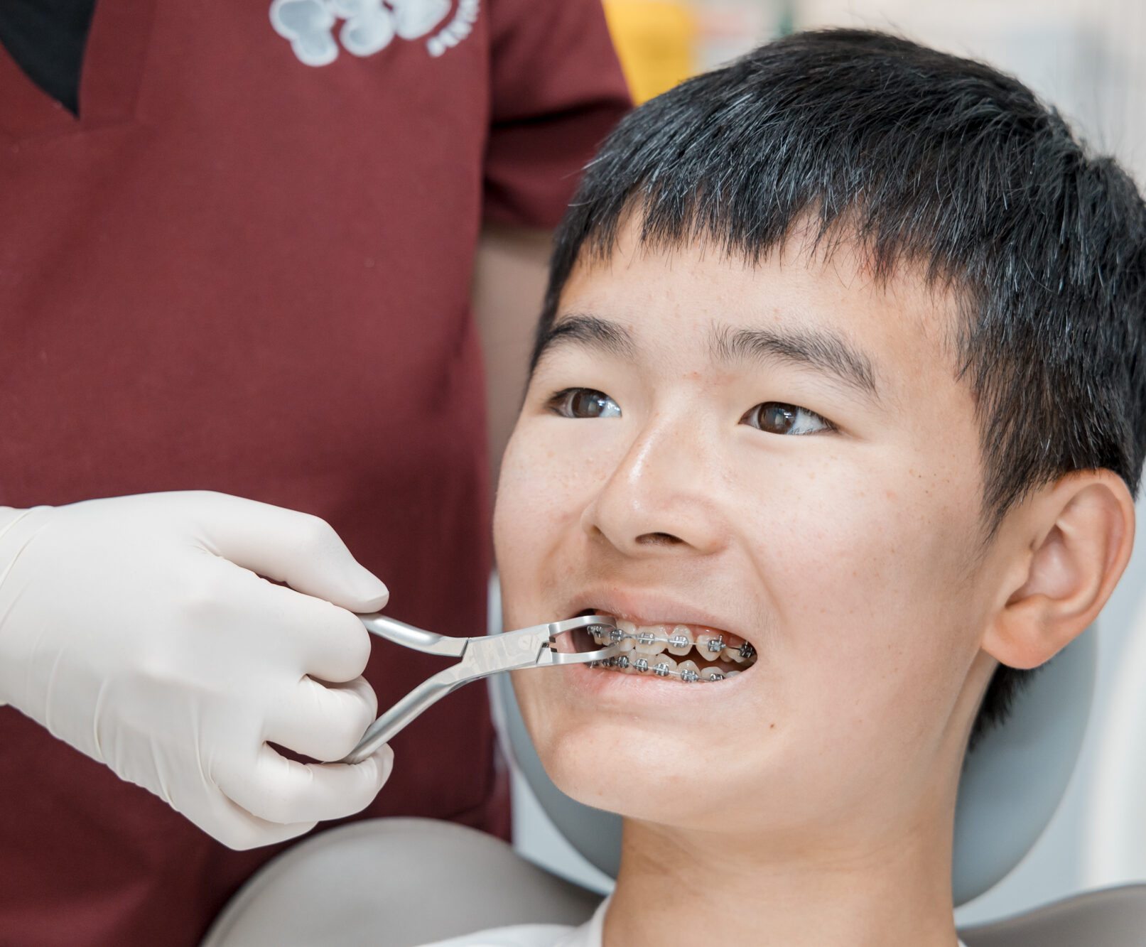 A dental professional in maroon scrubs adjusts a young patient's metal braces using a hand tool.