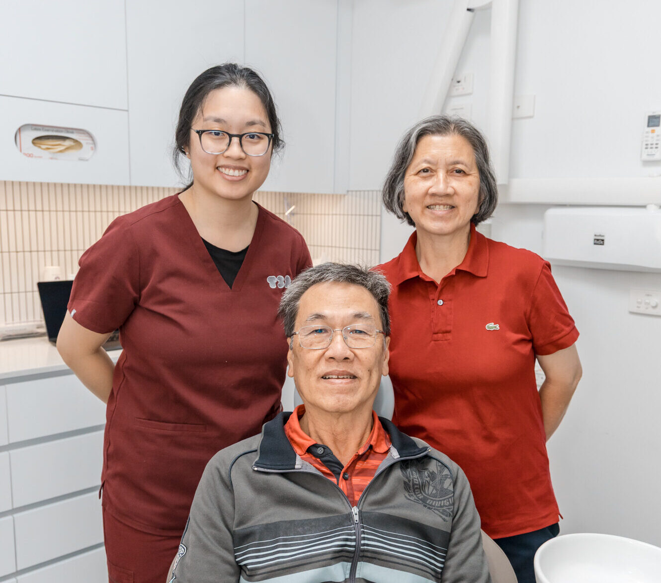 A happy dental implants patient smiles with comfort alongside his dentist and spouse in the clinic.