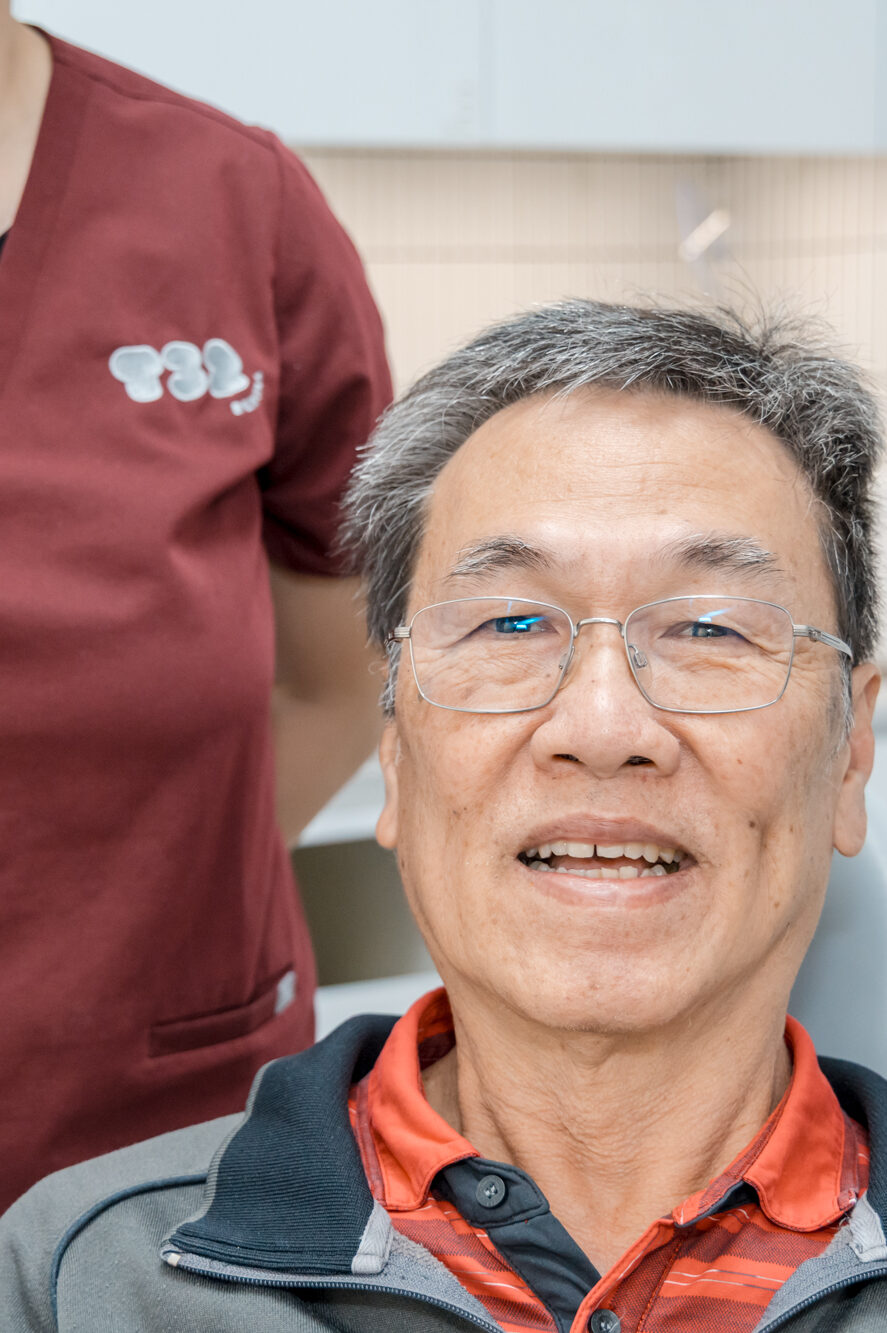 A smiling senior man with glasses sits in a dental chair with a clinical assistant in burgundy scrubs behind him.