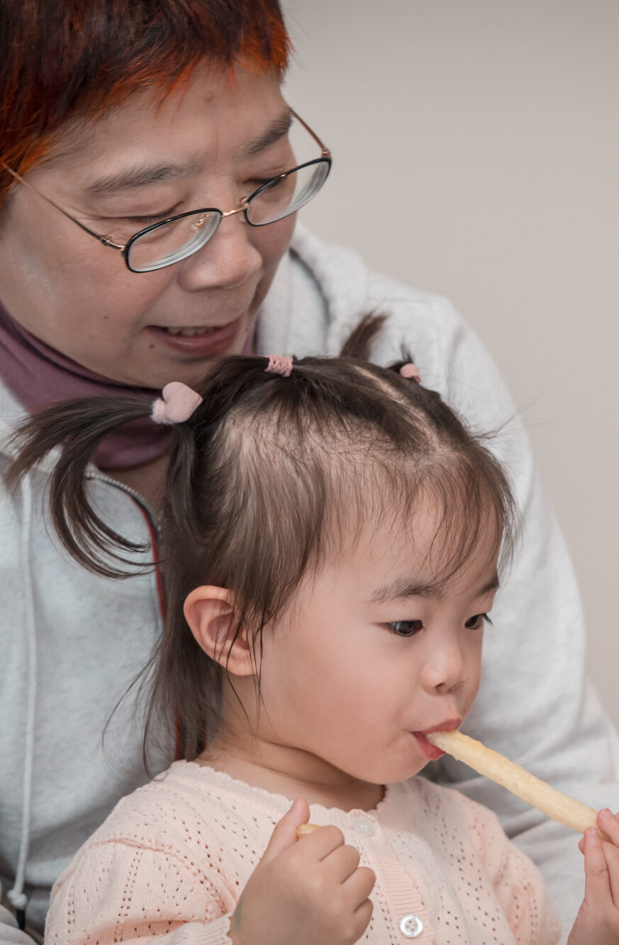 An older woman in glasses smiles while watching a toddler with pigtails eat a long, crunchy biscuit snack.