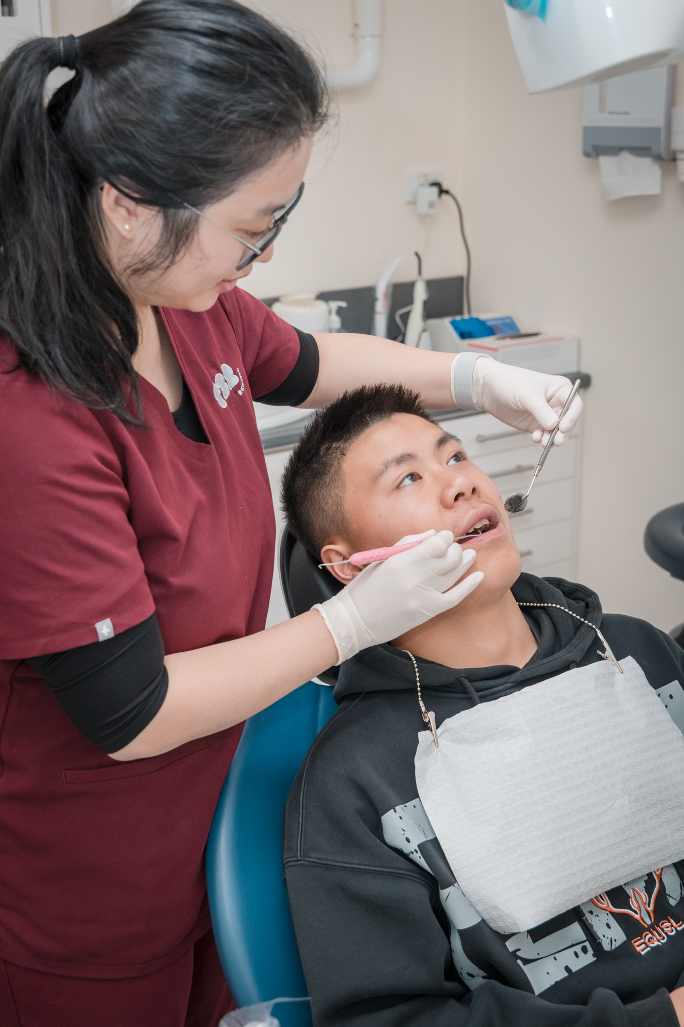 A dentist wearing red scrubs and gloves uses a dental mirror to examine a young male patient in a clinic.