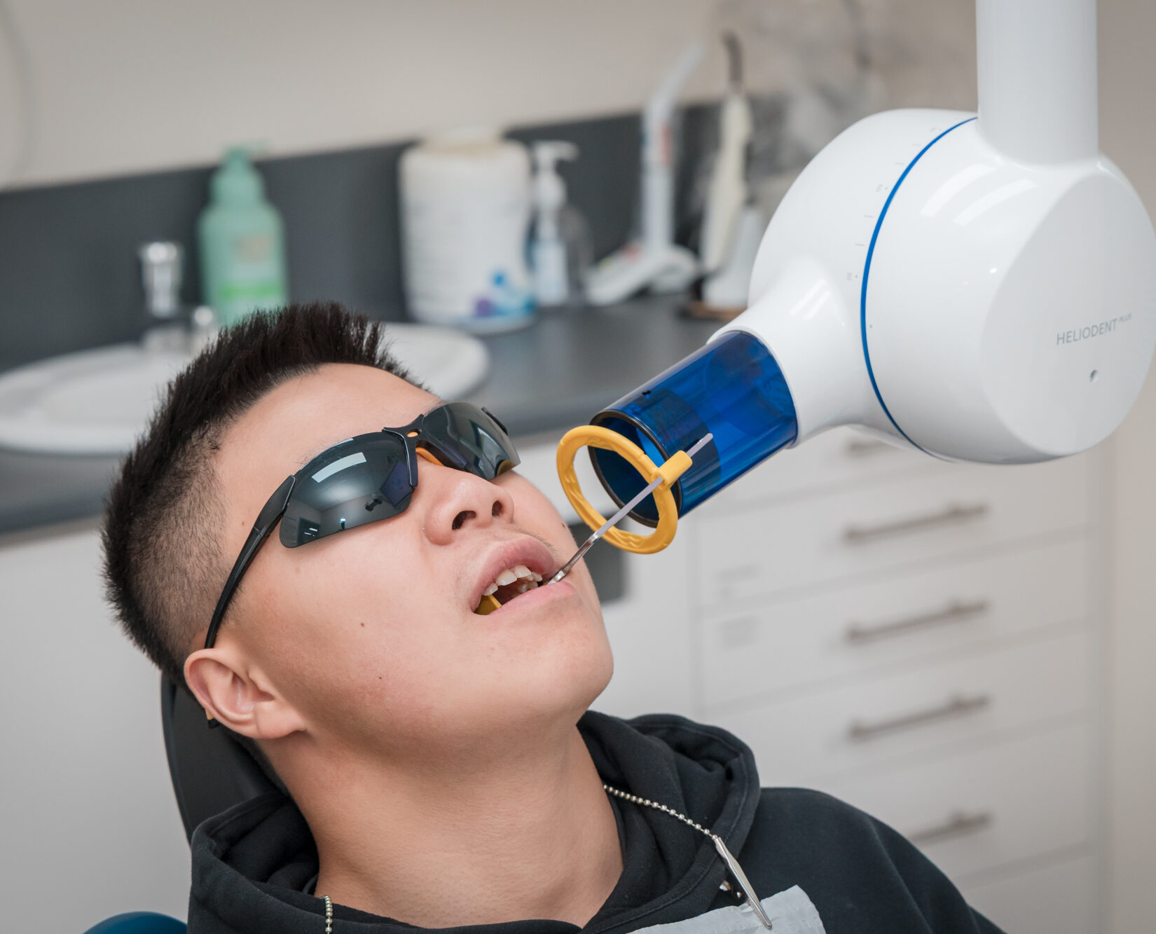 A patient wearing dark protective glasses lies in a dental chair as a white X-ray machine scans their mouth.