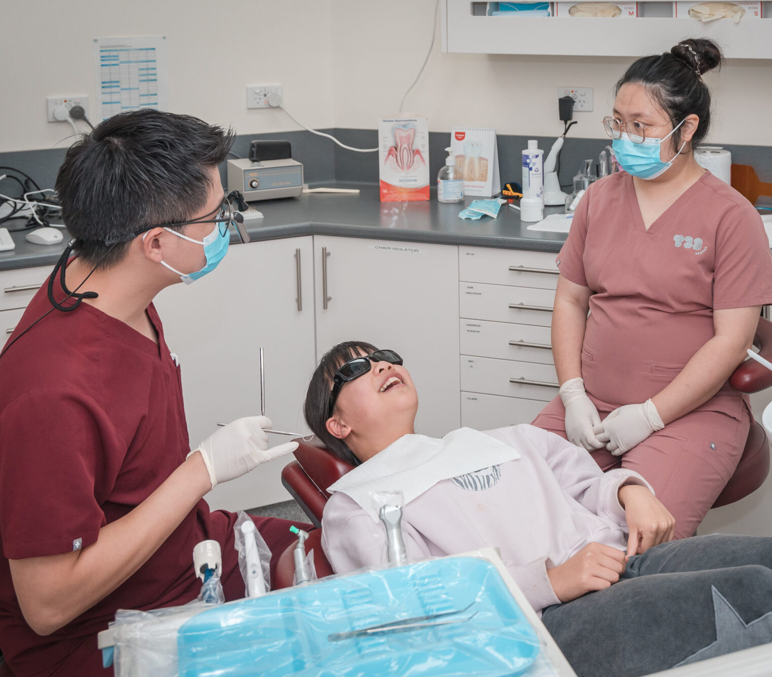 A male dentist and a dental assistant talk to a young patient wearing protective glasses in a dental chair.