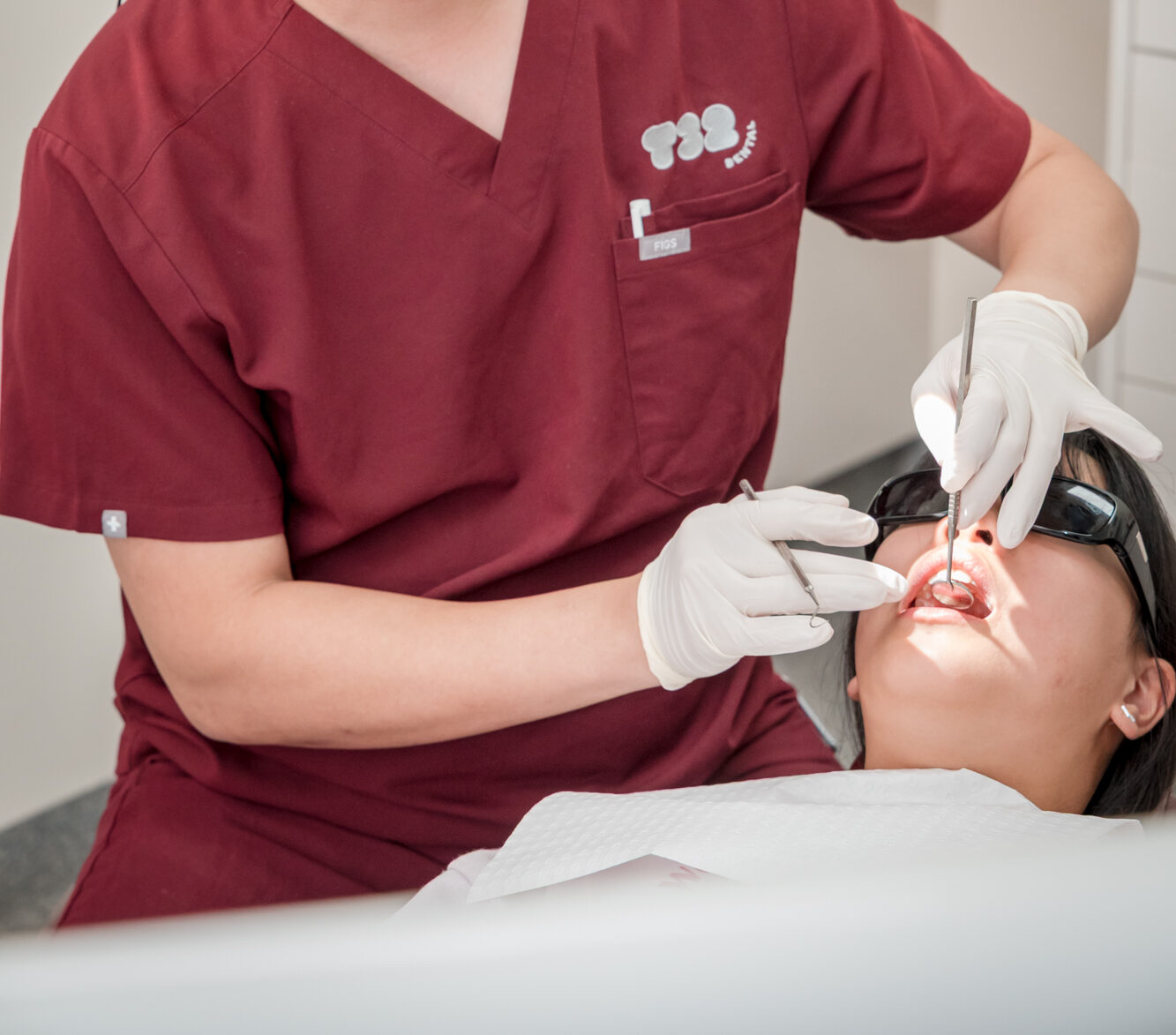 Dentist wearing maroon scrubs and magnifying glasses performs a clinical dental examination on a patient.