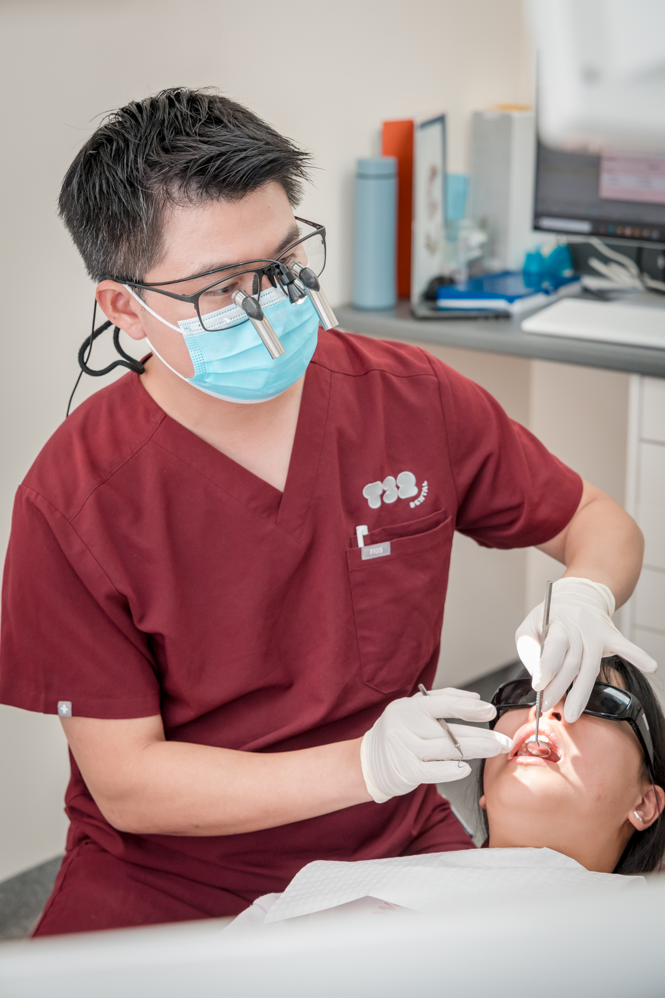 Dentist wearing maroon scrubs and magnifying glasses performs a clinical dental examination on a patient.