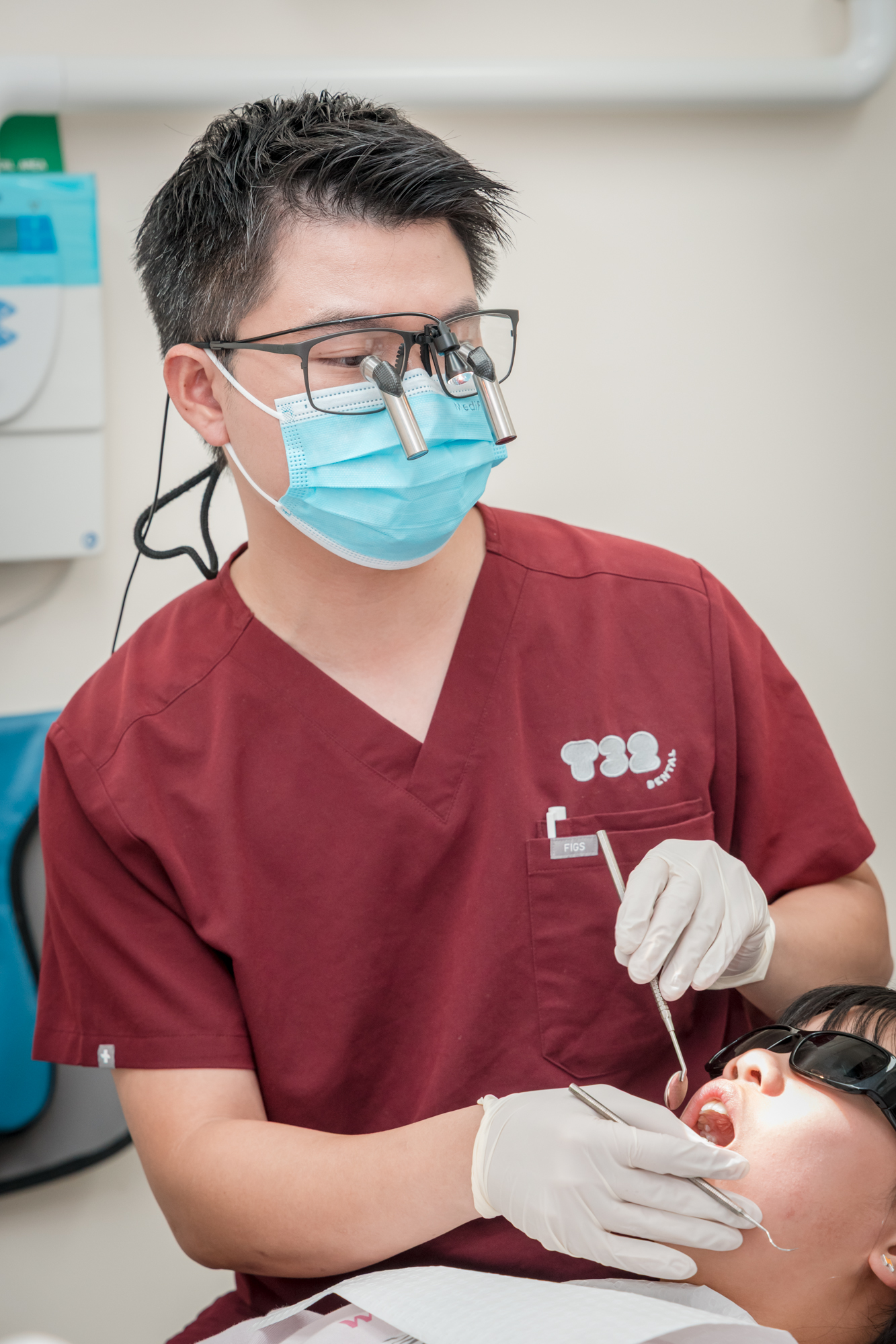 Dentist in maroon scrubs and magnifying loupes examines a patient's teeth with a dental mirror.