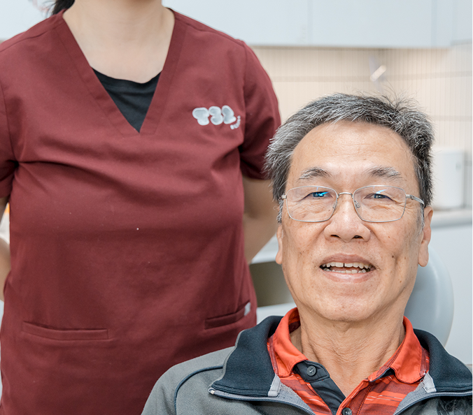 An older Asian man with glasses smiles in a dental chair with a clinical professional standing behind him.