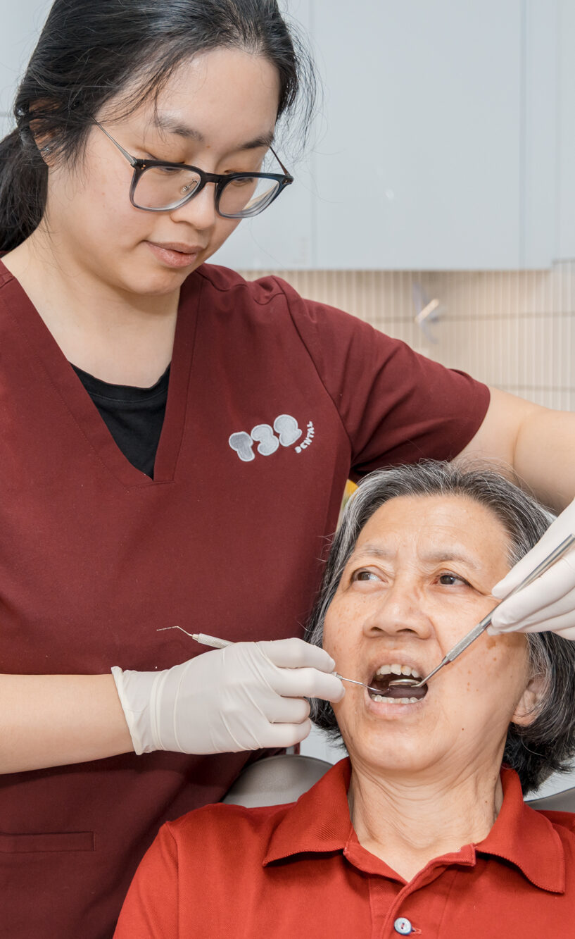 Dentist in maroon scrubs uses a dental mirror to examine an older patient's teeth during a checkup.