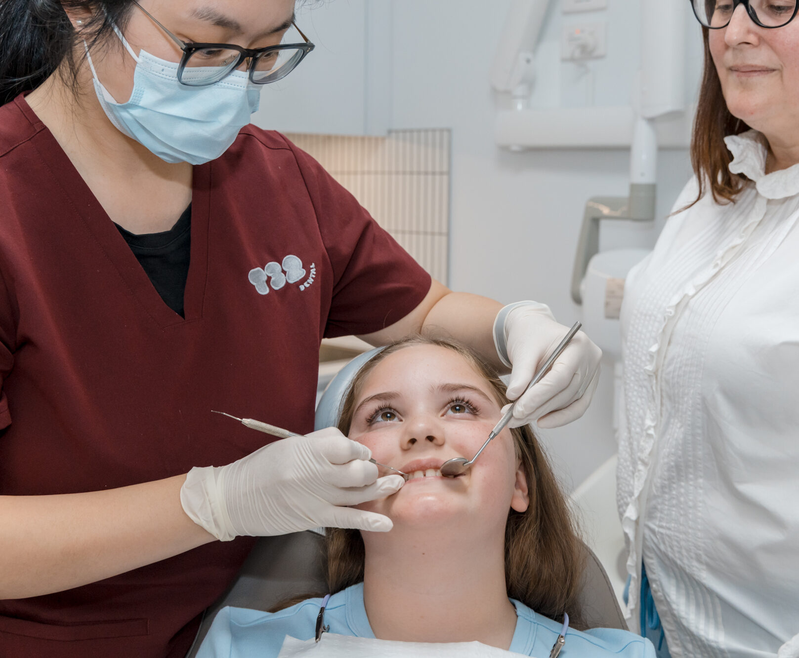 A female dentist in a mask and maroon scrubs examines a young girl's teeth while her mother watches.
