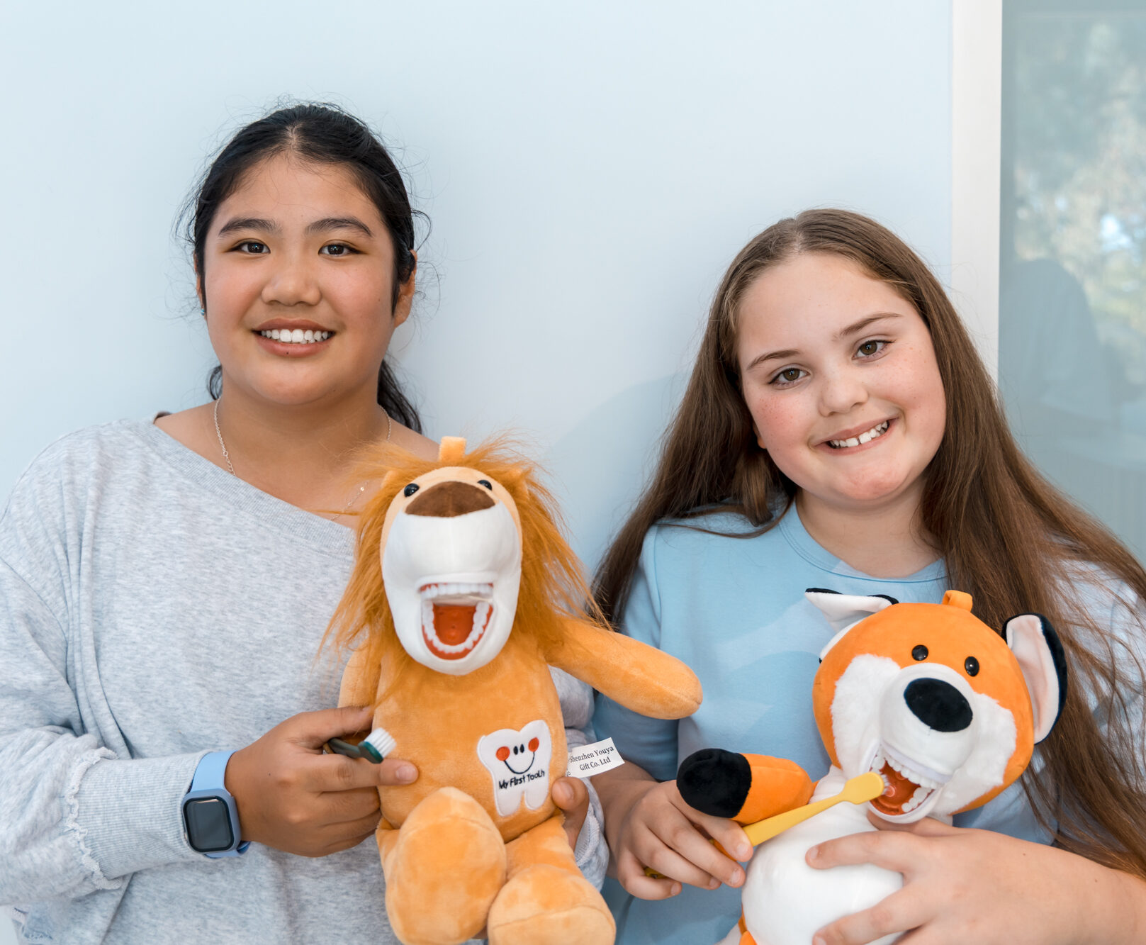 Two smiling girls holding plush lion and fox toys with human-like teeth and demonstrating brushing with toothbrushes.