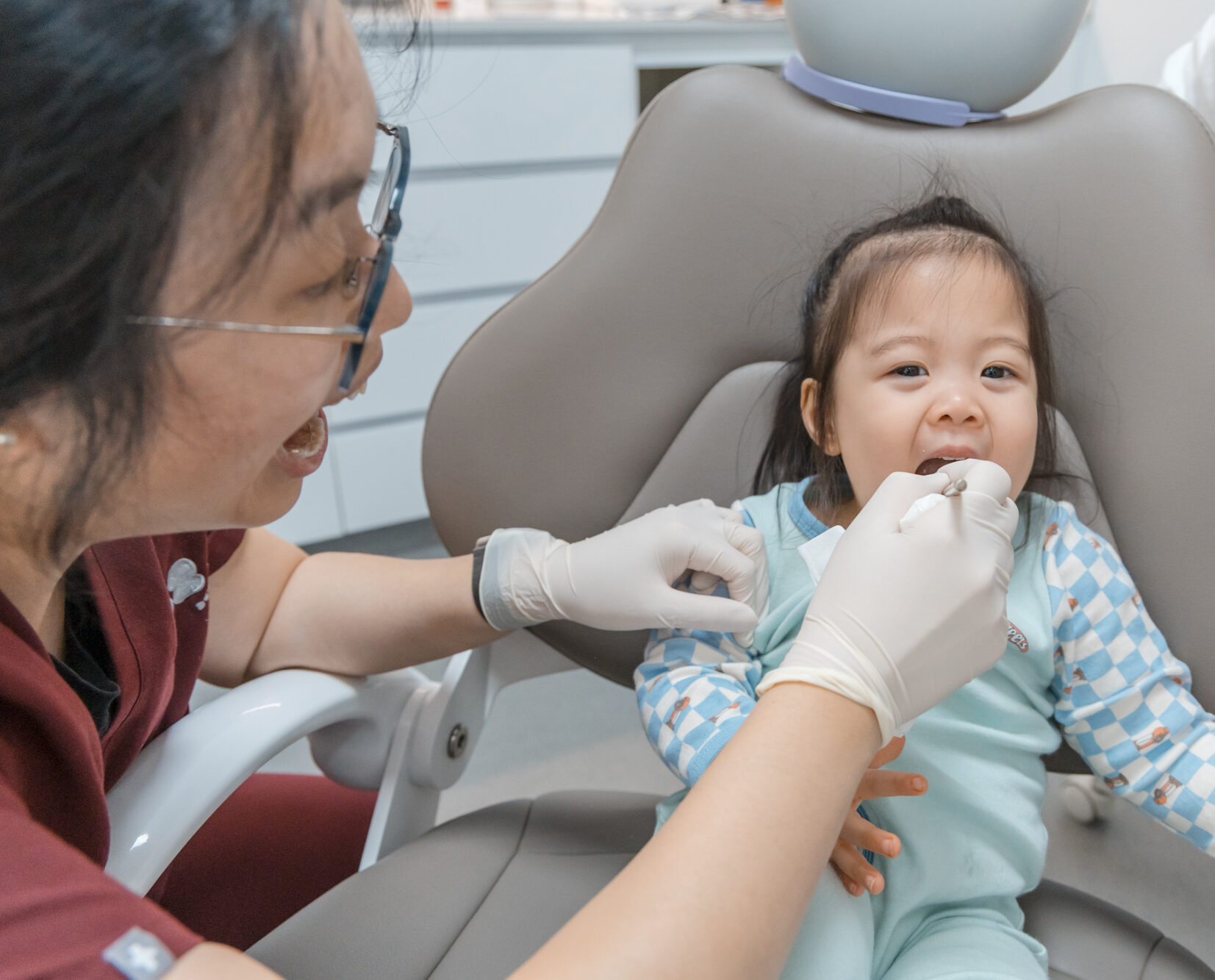 A cheerful toddler smiling during a gentle dental exam with a gloved dentist in a gray clinic chair.